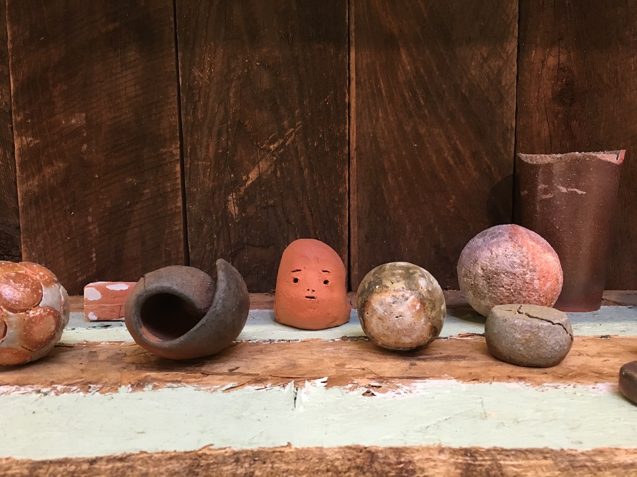 Roughly formed ceramic objects on a wooden surface. Spheres, a broken cup, and in the center a small unglazed piece with minimal markings of a face cut into it.
