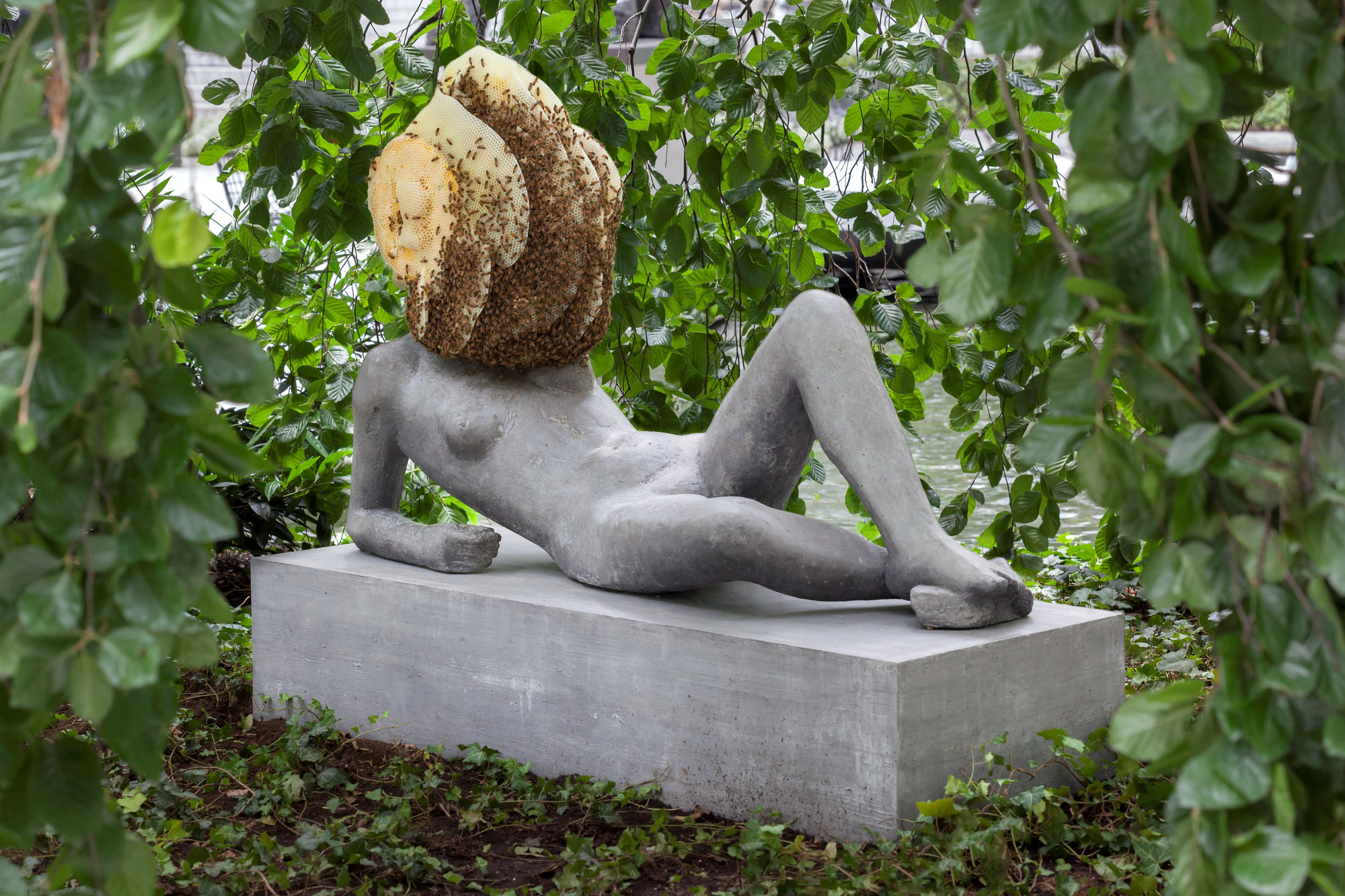 A stone statue of a nude reclining woman; in place of a head is a large, active bees nest.