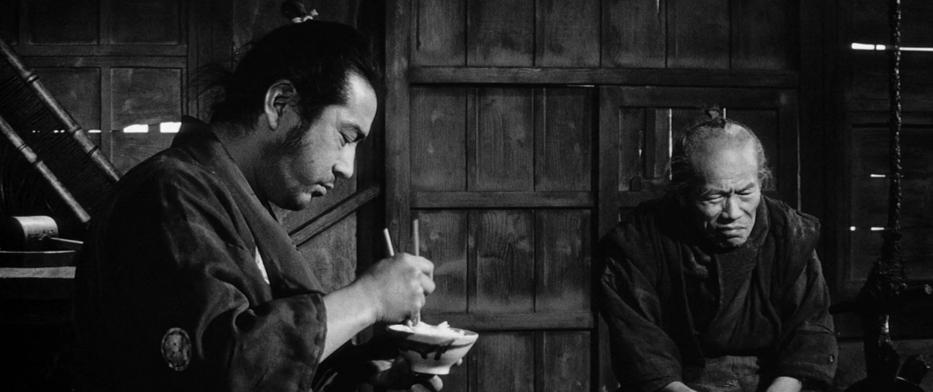 Black and white still from Akira Kurosawa samurai film. Toshiro Mifune eats rice out of a ceramic bowl with brush strokes; he is inside of a wooden shack, with an older man sitting to his right, scowling.