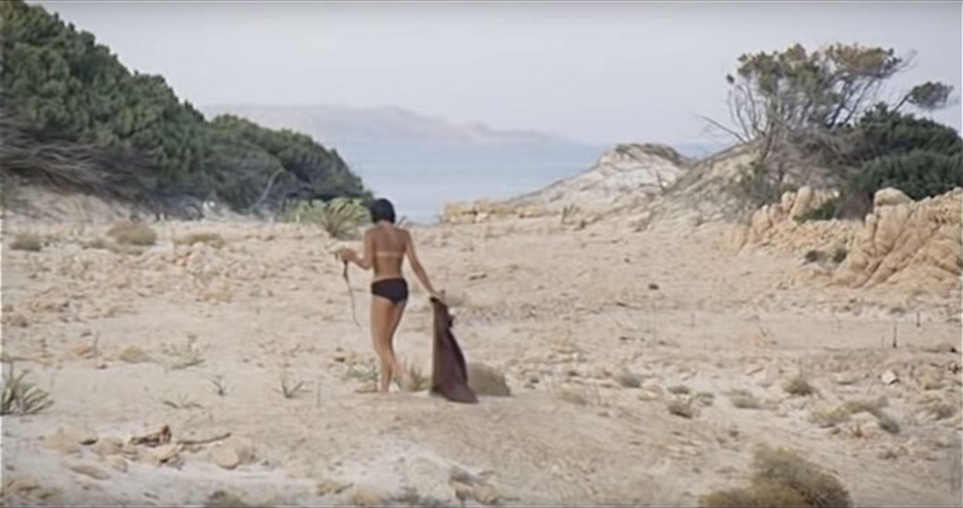 Still from a movie:
			Adolsescent girl wearing bikini bottom walking alone on a rocky beach, with a few desert plants in the background.