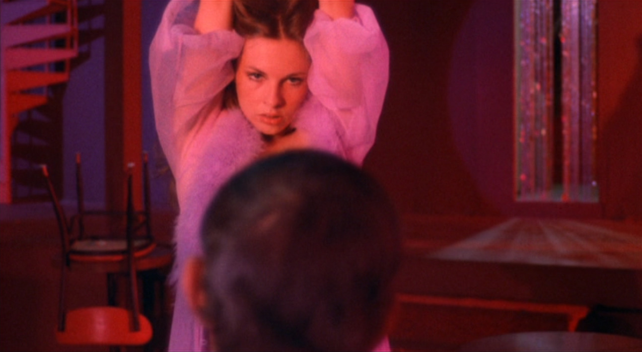 A still from a movie in which a woman gives a seductive look toward the camera. Her arms are held up above her head, and we can see the blurry back of a man's head in front of her, looking at her.