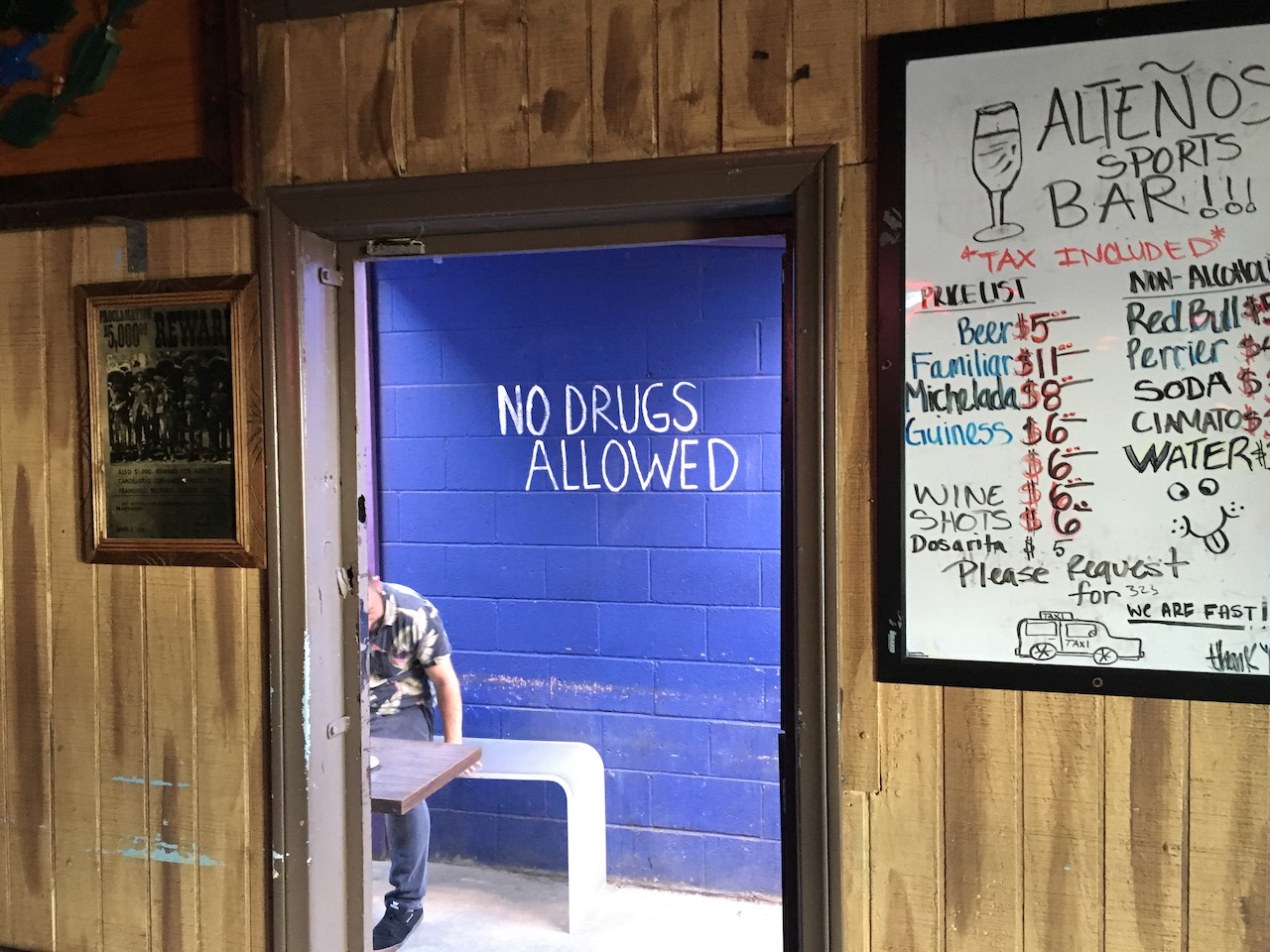 Photograph take inside a bar. On the wall a dry erase board with a drink menu 
        offers beer, wine, shots, etc. Through the door, a cinder block wall painted blue is visible, with the hand-painted 
        words NO DRUGS ALLOWED.