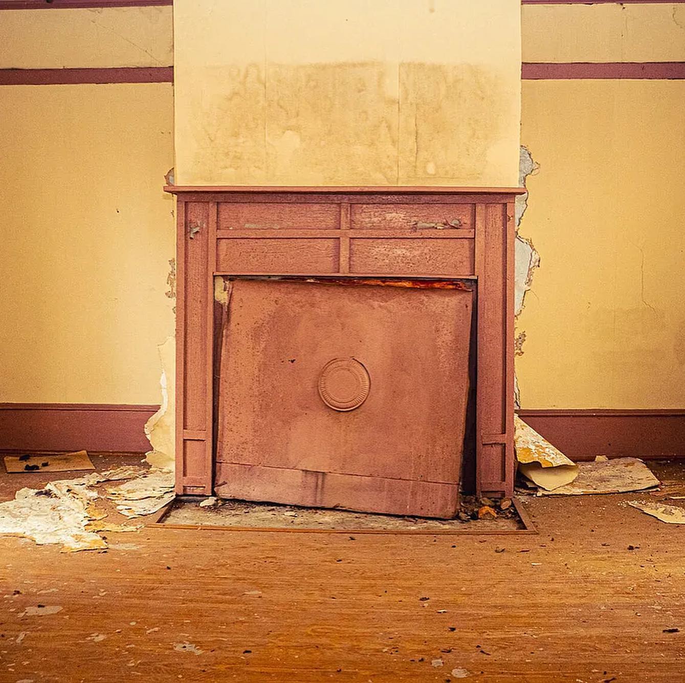 Photograph of the inside of an old house with no furniture. In the center is a fireplace, with aparent smoke stains above it. Plaster is falling down around it onto the wooden floor.