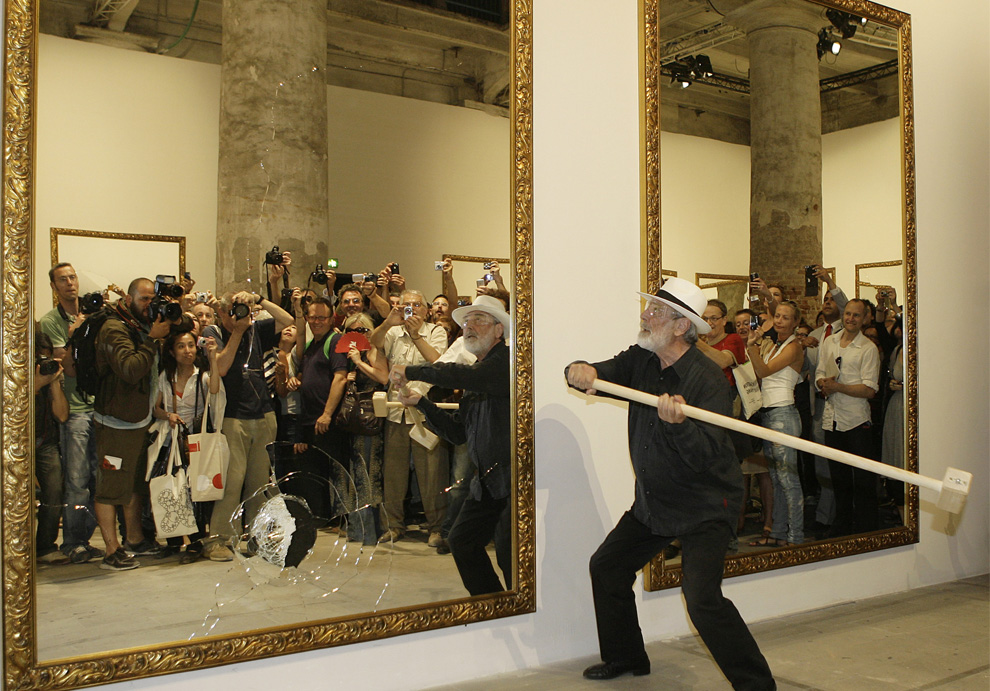 Older man wearing black clothes and a white hat, smashing a large mirror with a large mallet. In the mirror, a large group of people taking photographs can be seen behind him.