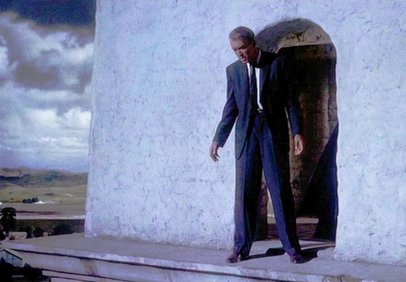 A man standing on the ledge of a bell tower looking down, an arched opening behind him