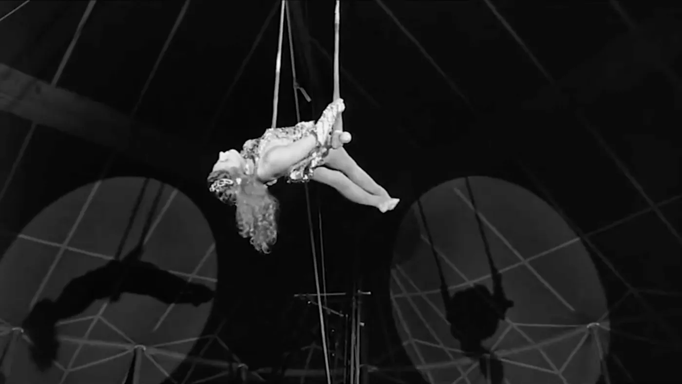 A still from Wings of Desire showing a trapeze artist. Her body is laying horizonally 
        with the trapeze across her back. Two spotlights shine from different directions creating 
        two shadows of her on the top of a circus tent behind her.