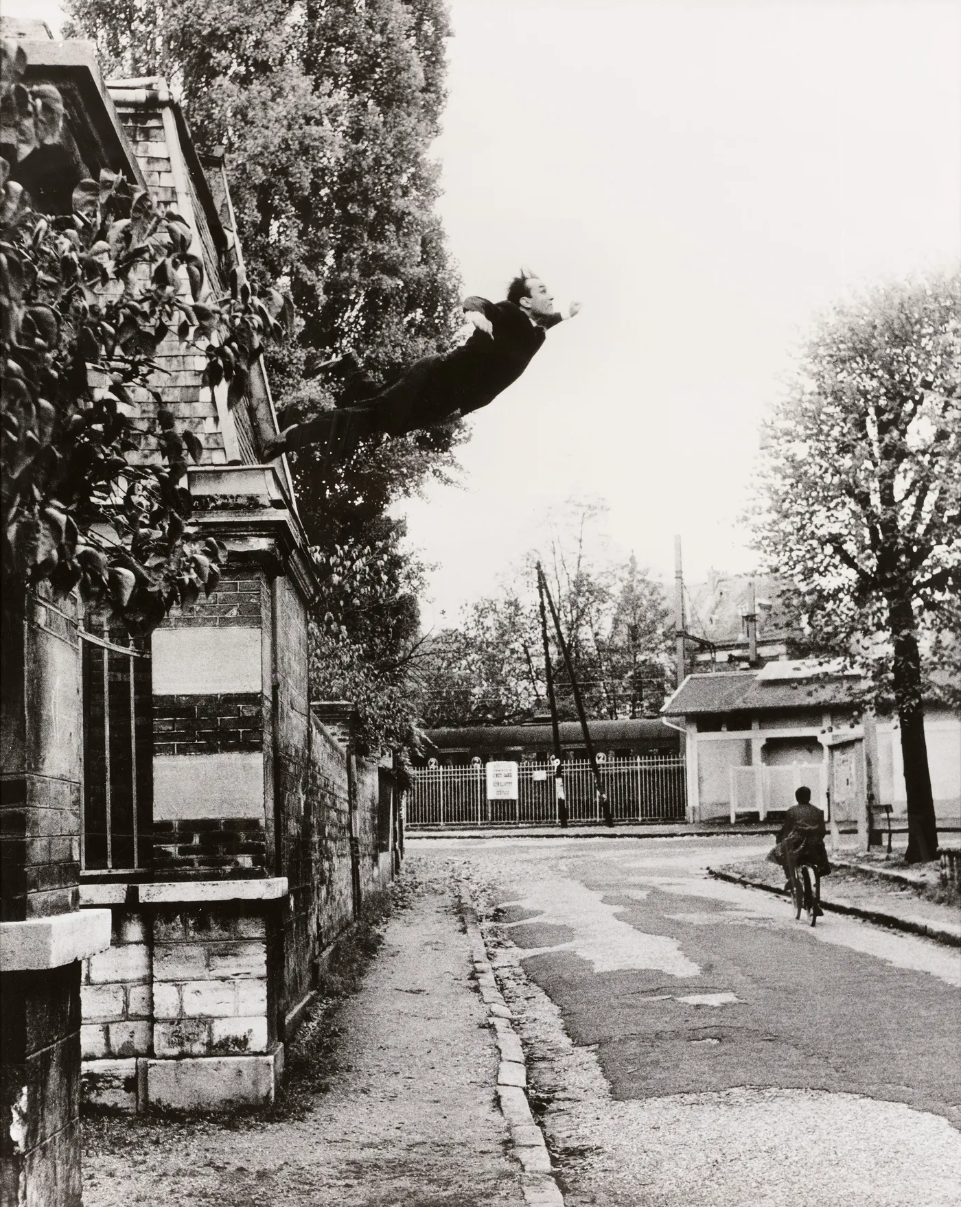 Black and white photograph of a man in mid-air leaping from a building wall over an empty street, arms outstretched